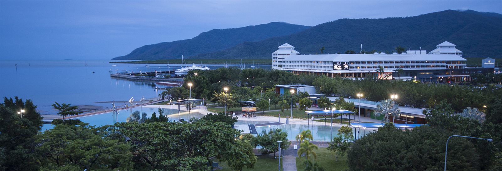 Cruise ship docked at a waterfront terminal beside a wooded hillside at dusk